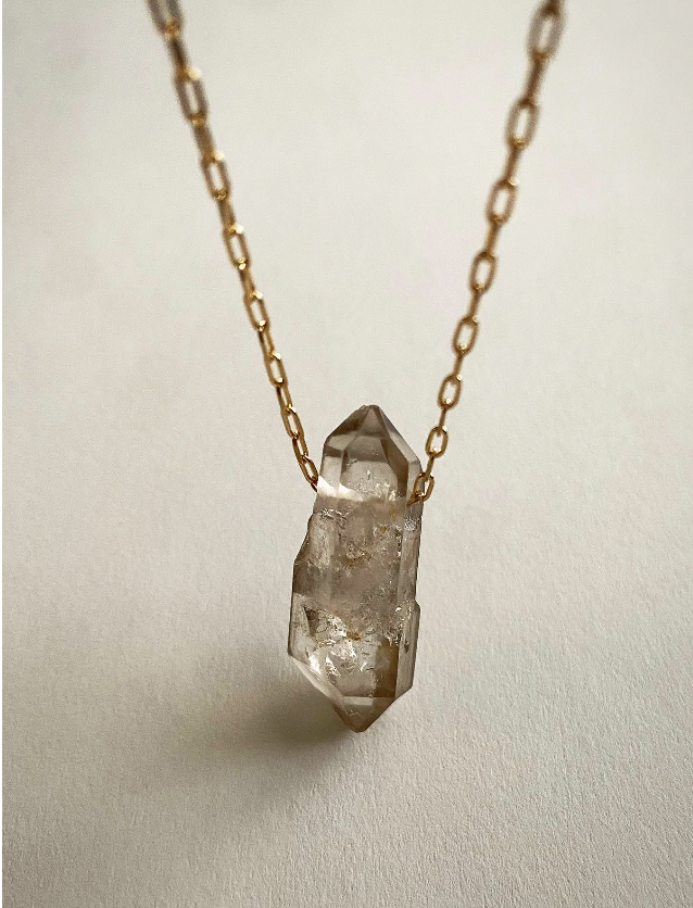 Raw clear quartz crystal pendant on a gold filled chain against soft cream linen